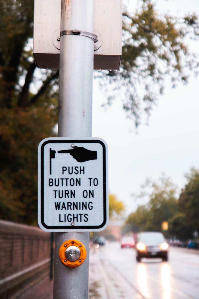Traffic sign instructing to push button to activate warning lights on a rainy street.